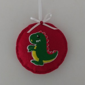 May include: A red felt ornament with a green and yellow dinosaur applique. The dinosaur has a red spine and is centered on the round ornament. A white ribbon is attached for hanging.
