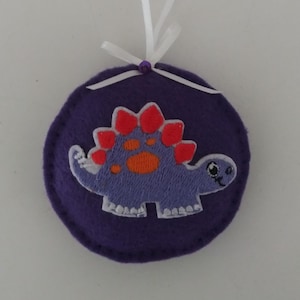 May include: A purple felt ornament featuring a dinosaur design. The dinosaur is light purple with orange and red accents. A white ribbon is attached for hanging. The ornament is round and has a stitched edge.