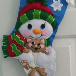 May include: A blue Christmas stocking with a snowman holding a cat. The snowman is wearing a green hat and a red scarf. The cat is brown and white.