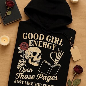 May include: Black hooded sweatshirt with the text "GOOD GIRL ENERGY" and a skull graphic holding a rose. The design includes a skeleton hand making an "okay" sign over an open book. The phrase "Open Those Pages JUST LIKE YOU SHOULD" is also printed on the front.