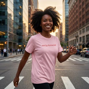 Könnte beinhalten: Hellrosa T-Shirt mit dem roten Schriftzug "easily distracted". Die Person, die es trägt, überquert eine Stadtstraße. Die Person lächelt und im Hintergrund sind Gebäude zu sehen.