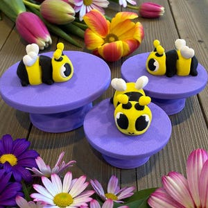 May include: A collection of whimsical bee-shaped figurines, each perched on a small, round, purple pedestal. The bees are yellow and black with white accents. The scene is decorated with colorful flowers, including tulips and daisies.