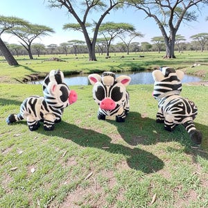 May include: Three plush zebra toys with black and white stripes, large black eyes, and pink noses are arranged on a grassy field. The zebras are in various poses, with a backdrop of trees and a small body of water under a clear blue sky.