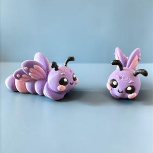 Puede incluir: Dos caprichosas figuras de mariposas moradas con antenas negras y mejillas rosadas. Una es una forma de oruga, la otra una mariposa estilizada. Las alas son un degradado de rosa y morado. Sobre un fondo azul claro.
