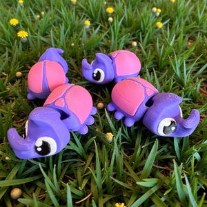 Könnte beinhalten: Mehrere lila und rosa Nashornfiguren mit großen Augen und gebogenen Hörnern. Sie sind auf einem Bett aus grünem Gras mit gelben Blumen angeordnet.