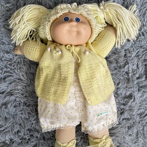 Könnte beinhalten: Eine Vintage-Cabbage-Patch-Kid-Puppe mit blonden Zopf-Zöpfen, blauen Augen und einem lächelnden Gesichtsausdruck. Die Puppe trägt eine gelbe Strickjacke, einen Strampler mit Blumenmuster und Spitzensaum sowie passende gelbe Strickschuhe. Die Puppe liegt auf einer grauen, flauschigen Oberfläche.