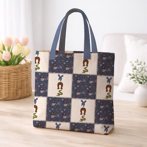 May include: A tote bag featuring a patchwork design of alternating squares. One set of squares displays a blue paisley pattern, while the other set features a white background with a brown horseshoe, a green snake, and a small blue bow motif.