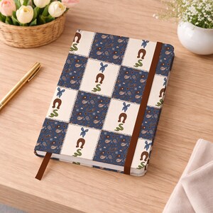 May include: A rectangular notebook with a checkered pattern featuring alternating squares of dark blue paisley print and cream white squares decorated with brown horseshoe icons, green squiggly lines, and small blue bows, secured by a brown elastic band.
