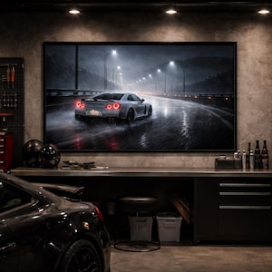 May include: A framed artwork depicting a silver sports car speeding down a wet road at night, illuminated by streetlights. The scene is set in a garage with tools, tires, and a black car in the foreground. The artwork is the focal point.