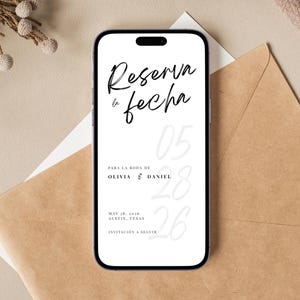 May include: A smartphone displays a wedding invitation with the text "Reserva la fecha" in elegant script. The invitation includes the names Olivia & Daniel, the date May 28, 2026, and the location Austin, Texas. The phone rests on a neutral-toned background.
