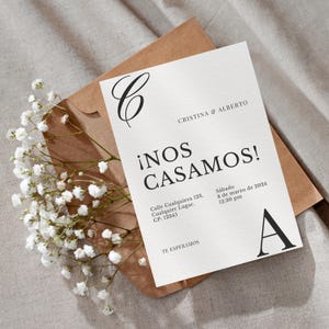 Peut inclure: Une invitation de mariage crème avec le texte noir "¡NOS CASAMOS!". L'invitation est posée sur une enveloppe marron, ornée de fleurs blanches. Les noms "CRISTINA & ALBERTO" et la date sont également présents.