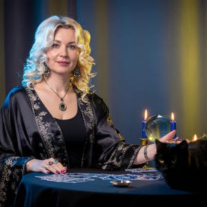 May include: A person with blonde hair wearing a black robe with gold floral patterns, a black top, and a pendant necklace. On the table are tarot cards, a clear crystal ball between two blue candles, and a black cat sitting near the edge.