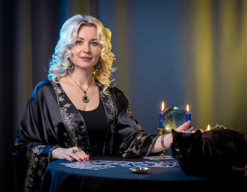 May include: A person with blonde hair wearing a black robe with gold floral patterns, a black top, and a pendant necklace. On the table are tarot cards, a clear crystal ball between two blue candles, and a black cat sitting near the edge.