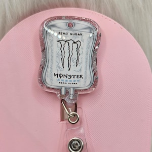 May include: A badge reel featuring a medical IV bag shape with a pink glitter resin finish. The centre displays the Monster Energy Zero Ultra logo and text, including the words Zero Sugar, Monster Energy, and Zero Ultra in black and white.