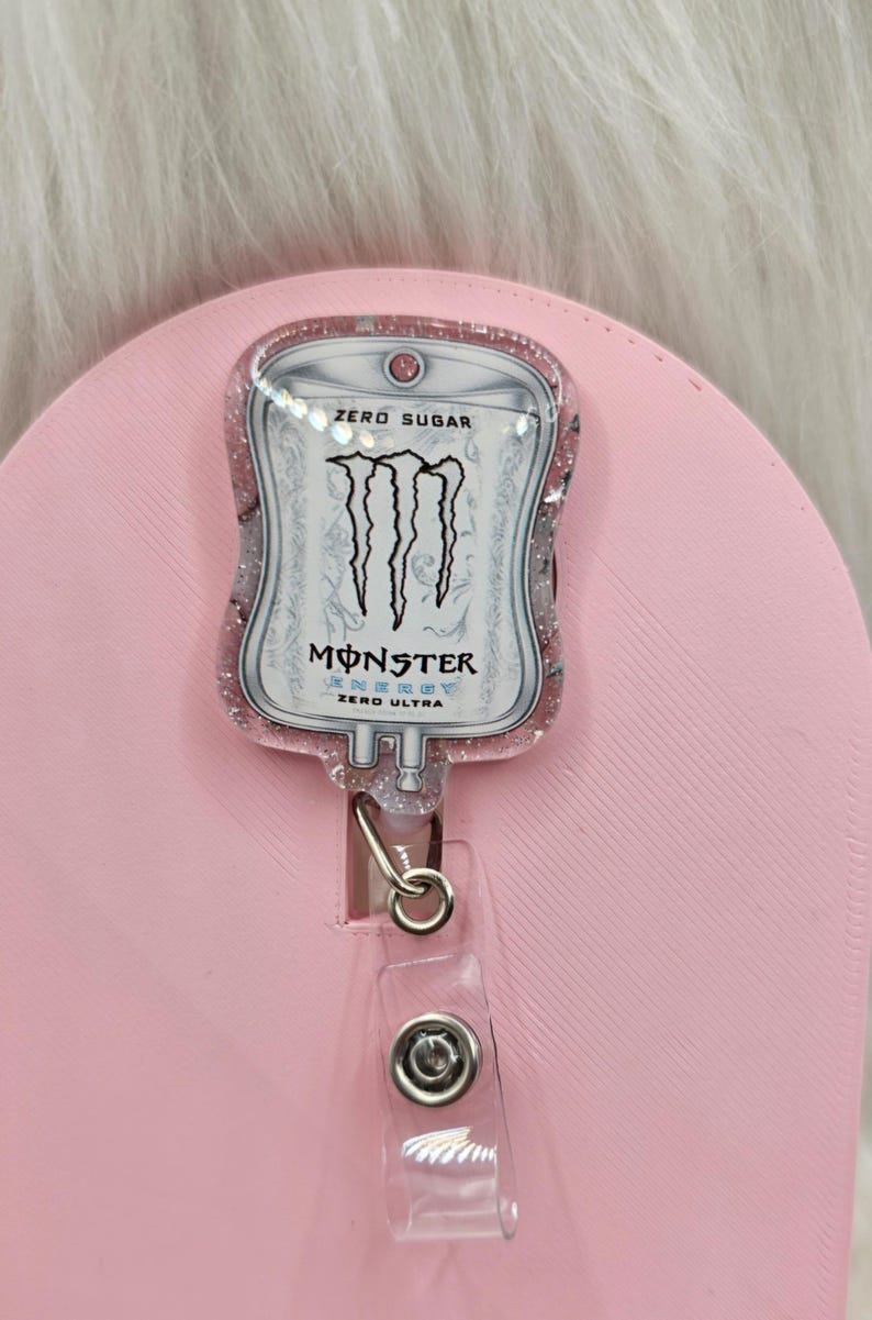 May include: A badge reel featuring a medical IV bag shape with a pink glitter resin finish. The centre displays the Monster Energy Zero Ultra logo and text, including the words Zero Sugar, Monster Energy, and Zero Ultra in black and white.