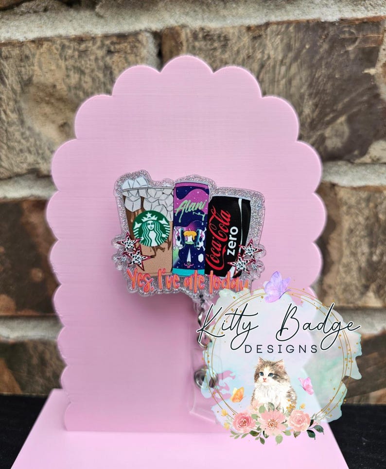 May include: An acrylic badge reel featuring a glittery design of three beverage containers: a Starbucks iced coffee, an Alani energy drink, and a Coca-Cola Zero can. The text "Yes, I've ate today" is printed in orange at the bottom of the graphic.