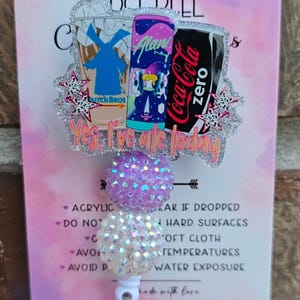 May include: A badge reel featuring a glittery acrylic topper with illustrations of a Dutch Bros cup, an Alani Nu energy drink, and a Coca-Cola Zero can. The text "Yes, I've ate today" is printed below the drinks. Two textured, iridescent beads sit below.