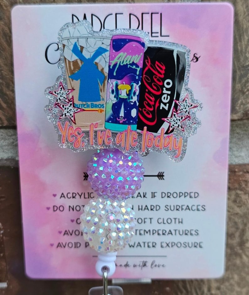 May include: A badge reel featuring a glittery acrylic topper with illustrations of a Dutch Bros cup, an Alani Nu energy drink, and a Coca-Cola Zero can. The text "Yes, I've ate today" is printed below the drinks. Two textured, iridescent beads sit below.