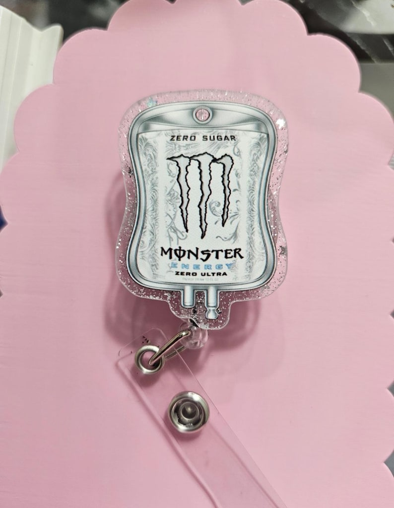 May include: A badge reel featuring a clear acrylic topper shaped like an IV bag with a Monster Energy Zero Ultra graphic. The design includes the black Monster logo, the text ZERO SUGAR, and ZERO ULTRA. The acrylic is filled with pink glitter.
