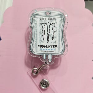 May include: A badge reel featuring a clear acrylic topper shaped like an IV bag with a Monster Energy Zero Ultra graphic. The design includes the black Monster logo, the text ZERO SUGAR, and ZERO ULTRA. The acrylic is filled with pink glitter.