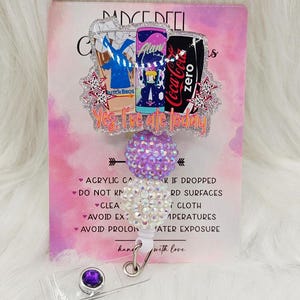 May include: A badge reel featuring a glittery acrylic topper with illustrations of a Dutch Bros cup, an Alani energy drink, and a Coca-Cola Zero can. The text reads, Yes, I've ate today. It includes a purple and a white rhinestone bead, and a clip.