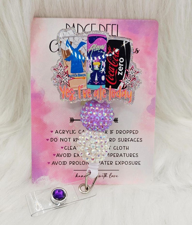 May include: A badge reel featuring a glittery acrylic topper with illustrations of a Dutch Bros cup, an Alani energy drink, and a Coca-Cola Zero can. The text reads, Yes, I've ate today. It includes a purple and a white rhinestone bead, and a clip.