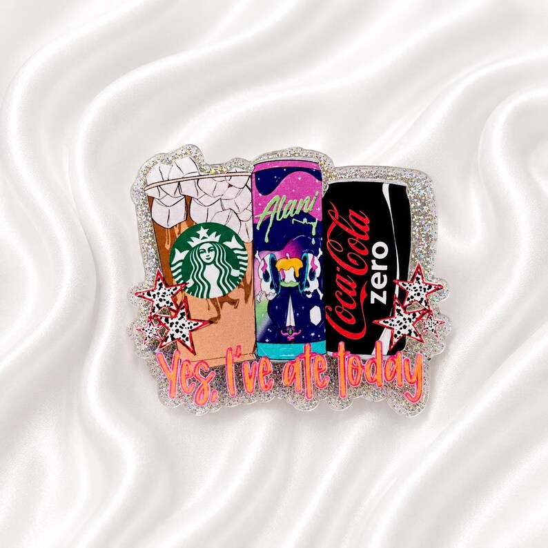 May include: A glittery acrylic badge featuring a Starbucks iced coffee, an Alani energy drink, and a Coca-Cola Zero can. The design includes small black and white star accents and the orange text Yes, I've ate today printed at the bottom.