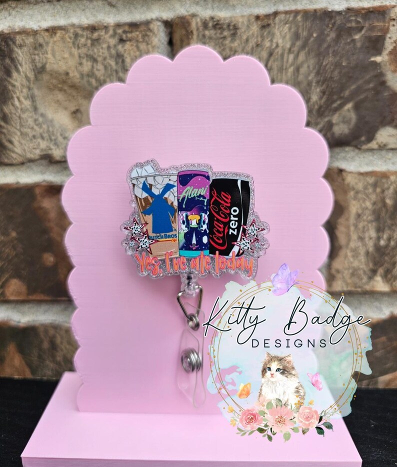 May include: An acrylic badge reel featuring a glittery design of three beverage containers, including a Dutch Bros cup, an Alani Nu can, and a Coca-Cola Zero can, with the text "Yes, I've ate today" printed in orange below the drinks.