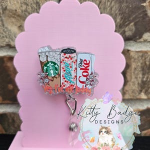 May include: A retractable badge reel featuring a clear acrylic topper with illustrations of a Starbucks iced coffee, an Alani Nu energy drink, and a Diet Coke can. The design includes small star accents and the orange text Yes, I've ate today.