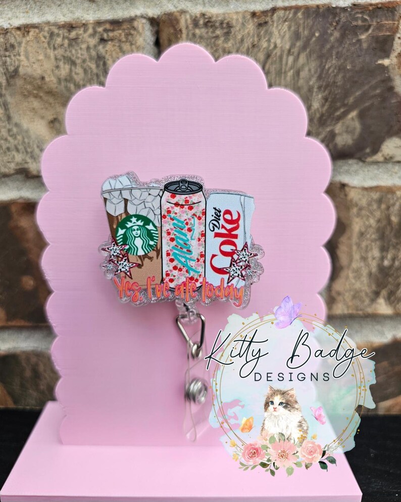 May include: A retractable badge reel featuring a clear acrylic topper with illustrations of a Starbucks iced coffee, an Alani Nu energy drink, and a Diet Coke can. The design includes small star accents and the orange text Yes, I've ate today.
