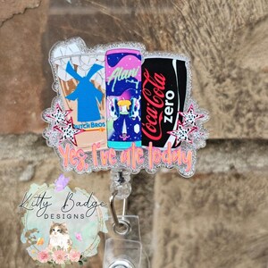 May include: A glittery acrylic badge reel featuring illustrations of a Dutch Bros cup, an Alani energy drink can, and a Coca-Cola Zero can. The design includes small star accents and the orange text Yes, I've ate today printed at the bottom.