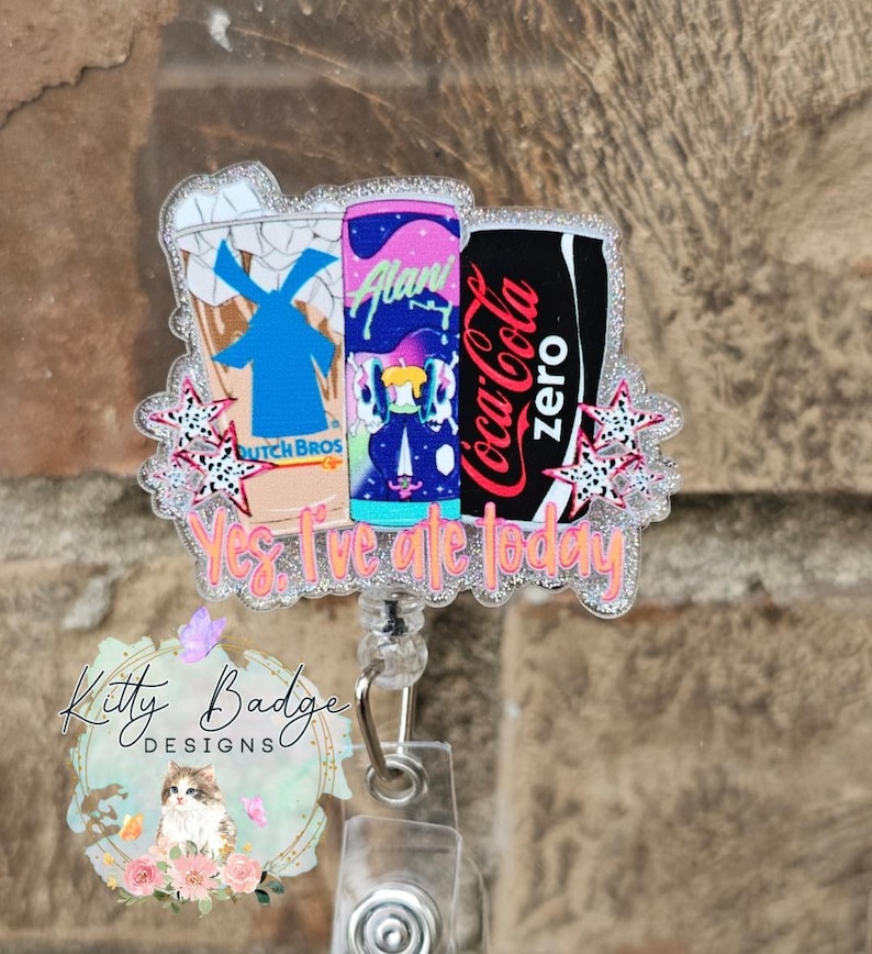 May include: A glittery acrylic badge reel featuring illustrations of a Dutch Bros cup, an Alani energy drink can, and a Coca-Cola Zero can. The design includes small star accents and the orange text Yes, I've ate today printed at the bottom.