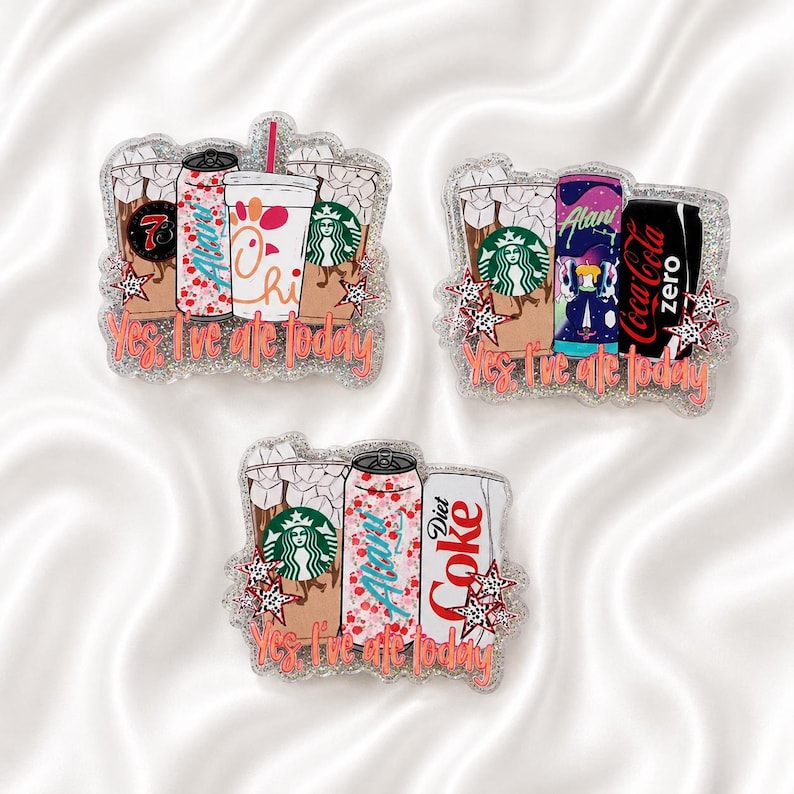 May include: Three glittery acrylic pins featuring illustrations of iced coffee cups, soda cans, and energy drinks. Each pin displays the text Yes, I've ate today in orange script, accented with small star shapes and various beverage brand logos.