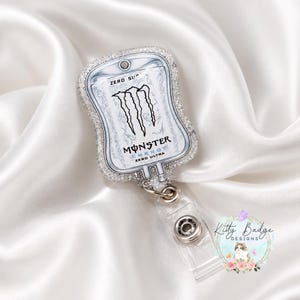 May include: A badge reel shaped like an IV bag featuring the Monster Energy Zero Ultra logo in black and white. The edges of the badge reel are covered in clear glitter, and it includes a clear plastic strap with a metal snap button closure.