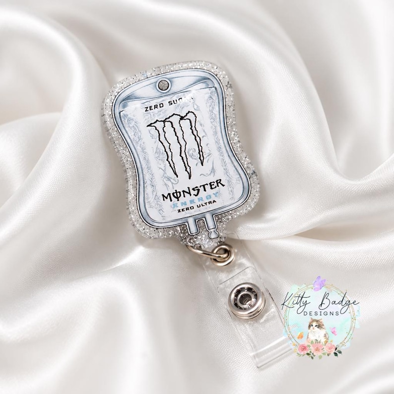 May include: A badge reel shaped like an IV bag featuring the Monster Energy Zero Ultra logo in black and white. The edges of the badge reel are covered in clear glitter, and it includes a clear plastic strap with a metal snap button closure.