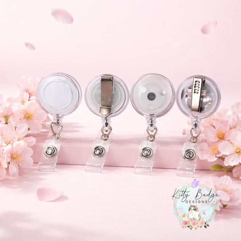 May include: Four circular badge reels with clear plastic housings and metal belt clips on the back. Each reel features a retractable cord attached to a clear vinyl strap with a metal snap button. The logo Kitty Badge Designs is visible.