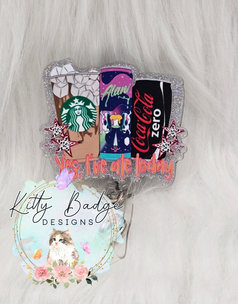 May include: A glittery badge reel featuring a Starbucks iced coffee cup, an Alani energy drink can, and a Coca-Cola Zero can. The design includes small star accents and the text Yes, I've ate today in orange lettering. The badge has a silver glitter border.