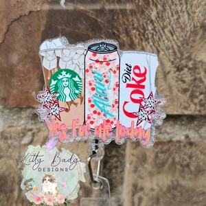 May include: A glittery acrylic badge reel featuring illustrations of an iced coffee cup with a green logo, a floral Alani can, and a Diet Coke can. The design includes small star accents and the orange text Yes, I've ate today.