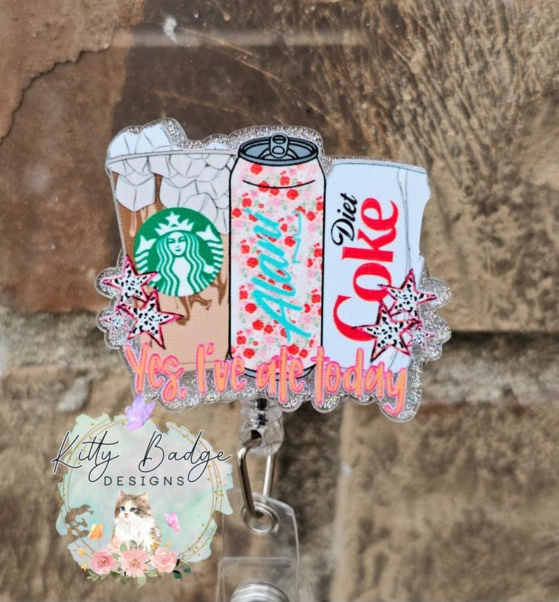 May include: A glittery acrylic badge reel featuring illustrations of an iced coffee cup with a green logo, a floral Alani can, and a Diet Coke can. The design includes small star accents and the orange text Yes, I've ate today.