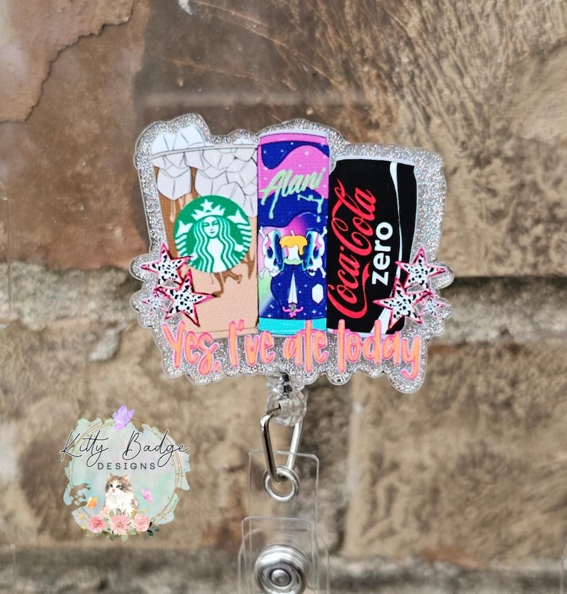 May include: A glittery acrylic badge reel featuring three beverage containers: a Starbucks iced coffee, an Alani energy drink, and a Coca-Cola Zero can. The design includes small pink star accents and the text Yes, I've ate today in orange.