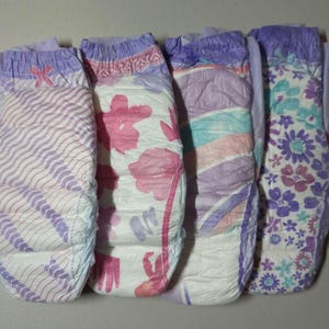 May include: Four pull-up diapers with different floral and patterned designs. The diapers feature a mix of colors, including white, purple, pink, and blue. Each diaper has a different design, including flowers and wavy lines.