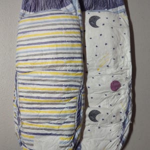 May include: Two baby diapers. One diaper has horizontal stripes in yellow, gray, and blue. The other diaper has a white background with a pattern of moons, stars, and a purple circle. Both diapers have blue and purple accents.