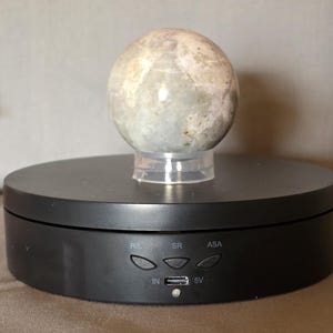 Puede incluir: Una piedra esf&eacute;rica de color claro se encuentra sobre un soporte de exhibici&oacute;n giratorio negro. La piedra tiene un aspecto moteado con tonos grises y beige. El soporte tiene botones de control etiquetados R/L, SR y ASA, junto con un puerto IN y una entrada de alimentaci&oacute;n de 5V.