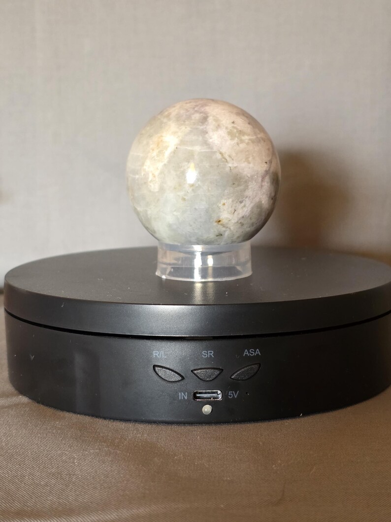 Puede incluir: Una piedra esf&eacute;rica de color claro se encuentra sobre un soporte de exhibici&oacute;n giratorio negro. La piedra tiene un aspecto moteado con tonos grises y beige. El soporte tiene botones de control etiquetados R/L, SR y ASA, junto con un puerto IN y una entrada de alimentaci&oacute;n de 5V.