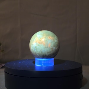 May include: A spherical, polished stone with a mottled green and gold surface. The stone sits atop a black, rotating display stand with a blue light illuminating the base. The background is a neutral, textured fabric.