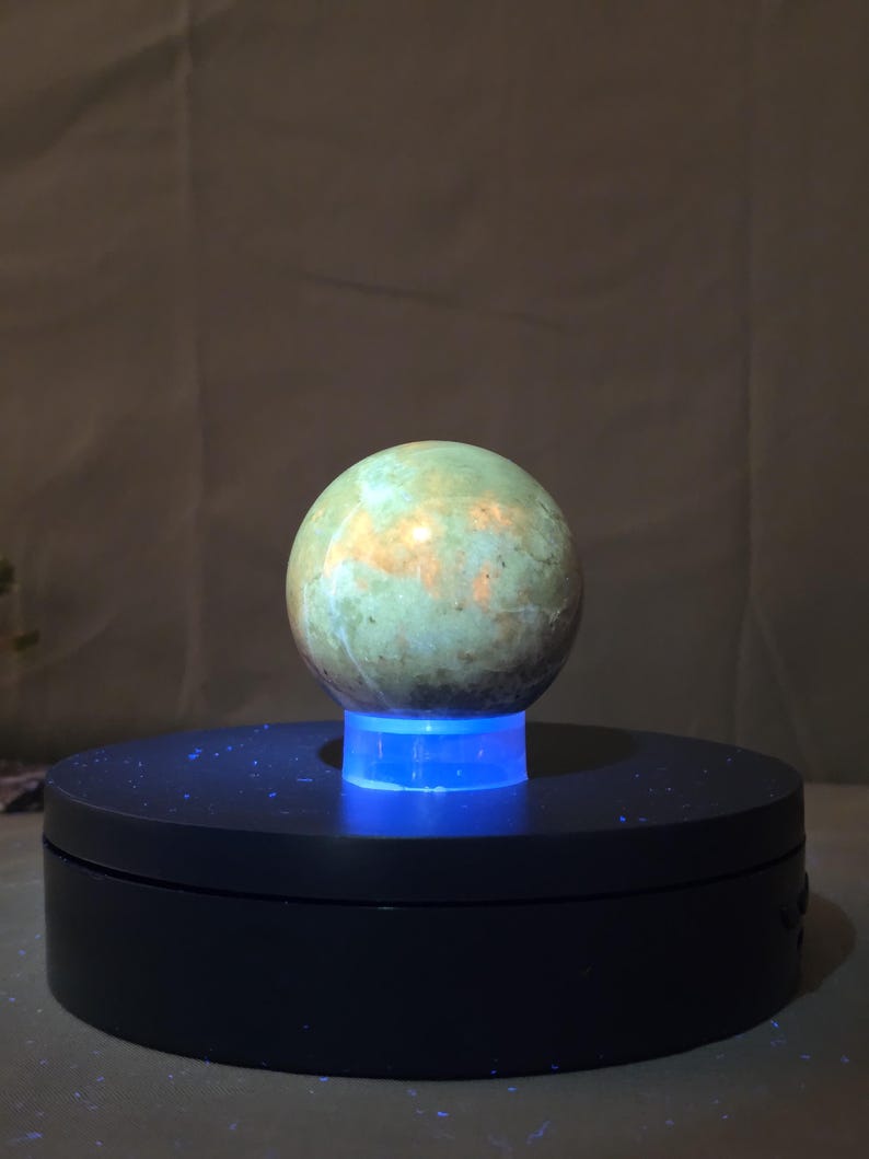 Puede incluir: Una piedra esf&eacute;rica y pulida con una superficie moteada verde y dorada. La piedra se encuentra sobre un soporte de exhibici&oacute;n giratorio negro con una luz azul que ilumina la base. El fondo es una tela neutra y texturizada.