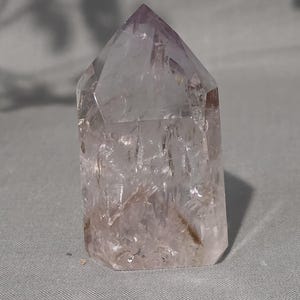 May include: A polished amethyst crystal point with a hexagonal base and a pointed top. The stone features a translucent, pale purple hue with visible internal fractures, natural inclusions, and clear crystalline facets throughout the structure.