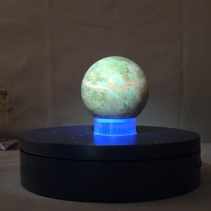 Puede incluir: Una esfera verde y naranja, posiblemente una gema, se encuentra sobre una plataforma iluminada en azul. La esfera tiene una superficie texturizada con vetas naranjas y azules. La plataforma est&aacute; sobre una base circular negra.