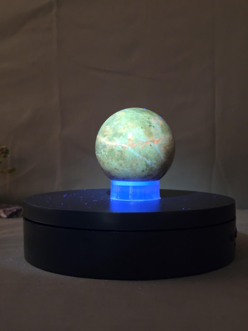Puede incluir: Una esfera verde y naranja, posiblemente una gema, se encuentra sobre una plataforma iluminada en azul. La esfera tiene una superficie texturizada con vetas naranjas y azules. La plataforma est&aacute; sobre una base circular negra.