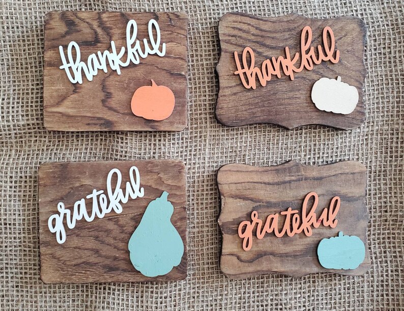 May include: Four wooden signs with the words "thankful" and "grateful" written in white and orange paint. Each sign has a cutout of a pumpkin or pear in orange, white, or blue.